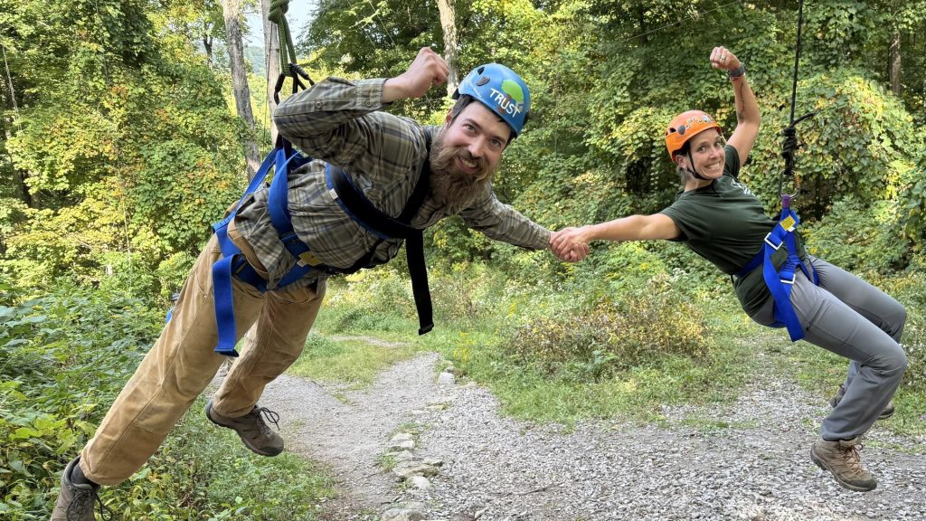 Two people ziplining through a forest, surrounded by tall trees and greenery.